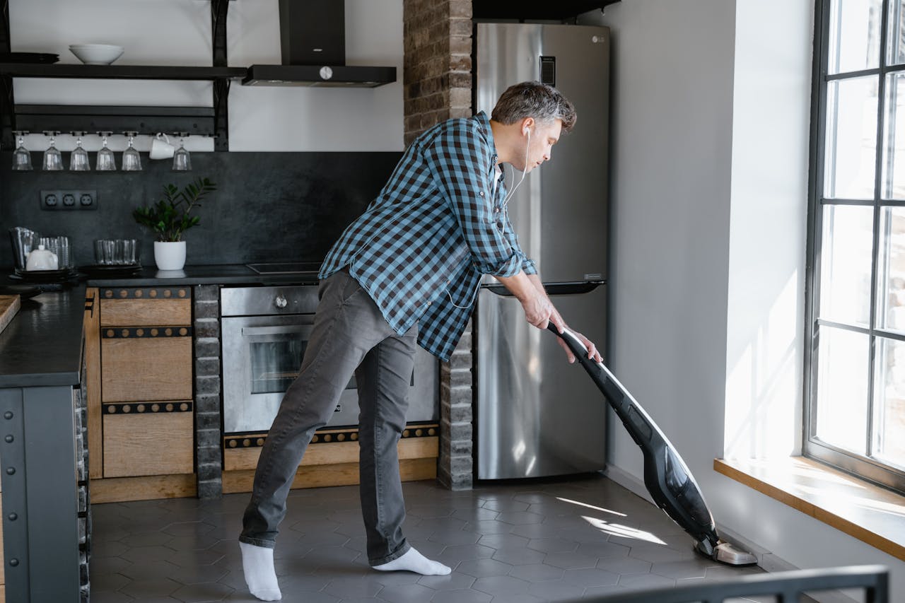 Adult man wearing earphones while vacuuming the kitchen floor at home, adding a modern lifestyle twist to household chores.